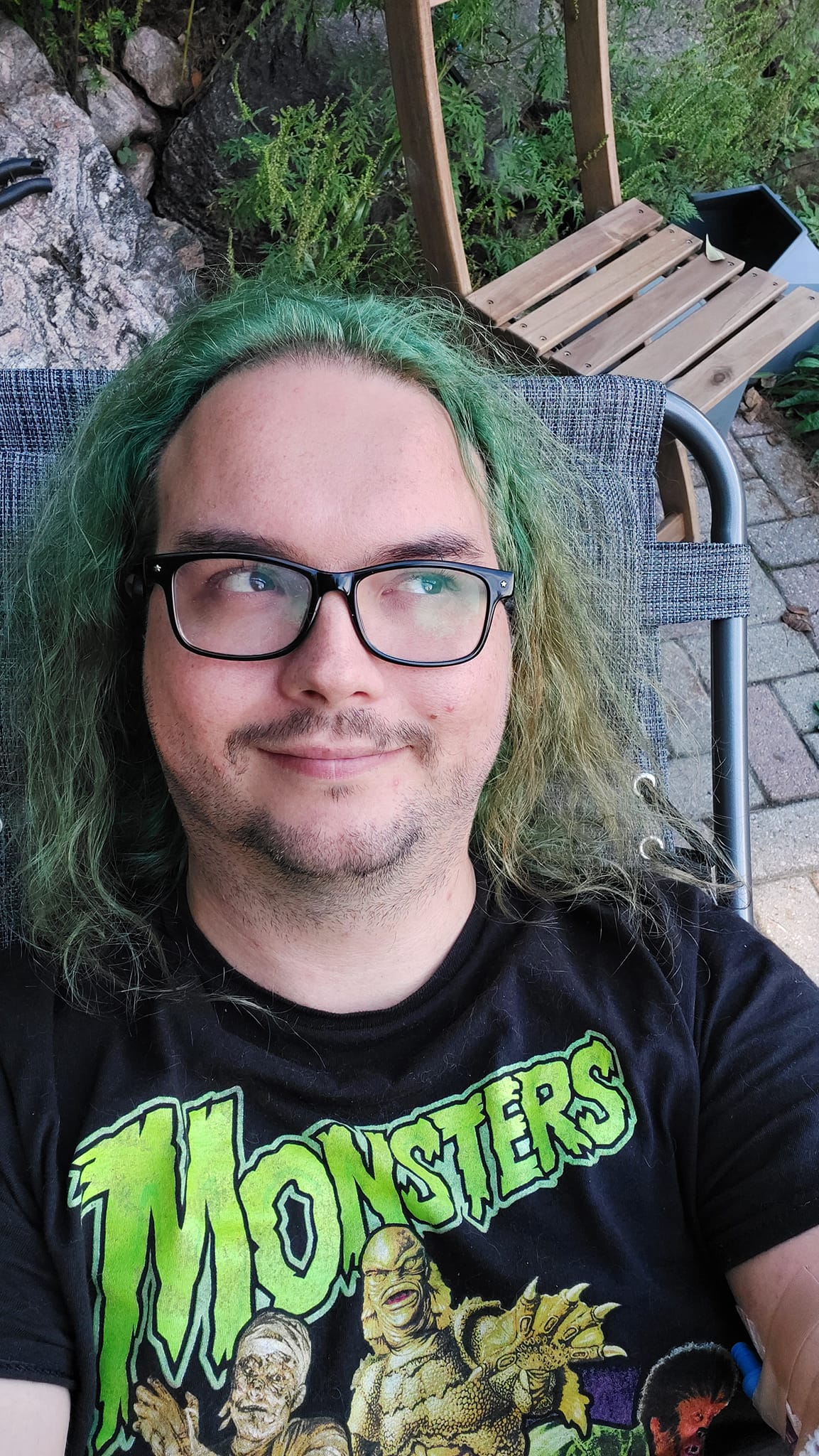 A man with long green hair and no beard sits lays in a folding chair on a patio, Photo 2